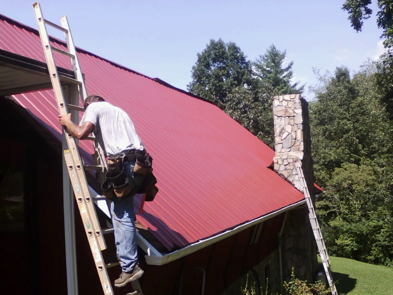 Roofer on a ladder beside a red metal roof and stone chimney — Soffit & Fascia Repair by Roofing Brothers in Cranberry